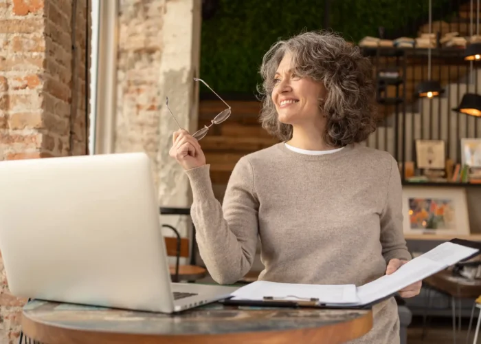 Mulher idosa sorrindo com cabelos cacheados, usando um suéter cinza claro, segurando óculos e com um caderno aberto na mesa, trabalhando em um ambiente de escritório com fundo de parede de tijolos e prateleiras de livros.